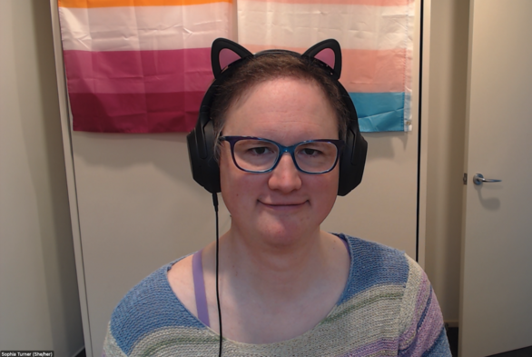 Me with a cute top, glasses, cat ears on my headphones, and both trans pride and lesbian pride flags in background