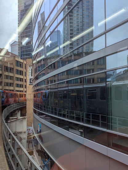 Photo taken from inside a commuter train that is going around a curve on an elevated train track. The front of the train is visible on the left, but most of the train is reflected in the glass of the building that the train is curving around.