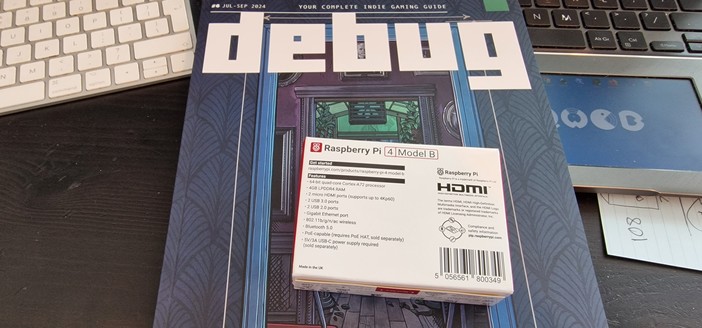 Photo of a Raspberry Pi on top of Debug magazine issue 6. My desk with keyboards and laptops in the background.