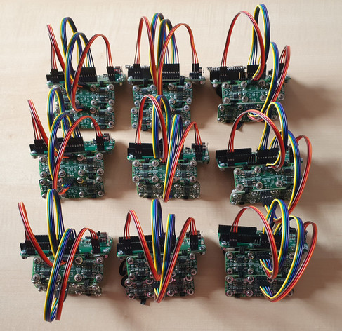 A photograph of 9 PsyLink devices, which are armbands with electrodes for remote-controlling computers with arm gestures. They're 3 green circuit boards connected with rainbow-colored wires.