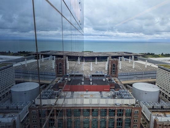 A view from a high rise window looking onto buildings and a large body of water fading from turquoise to blue meeting a gray blue fluffy clouds at the  horizon. The left half of the photo is a mirror of the right, reflected on the glass window facade of the building. 