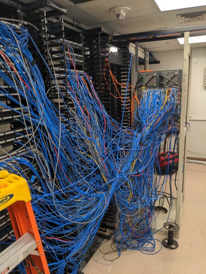 A set data racks with a jumbled mess of tangled patch cables streched to a different rack containing several switches and pulled out into the room. The cables are piled twisted and stretched all which way.