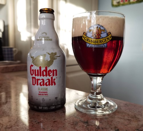 A stubby bottle of Gulden Draak in the famous white color next to the beer poured into a Grimbergen glass. Both are sitting on a stone countertop. The beer is reddish-brown in color with a large head of reddish-tan foam.