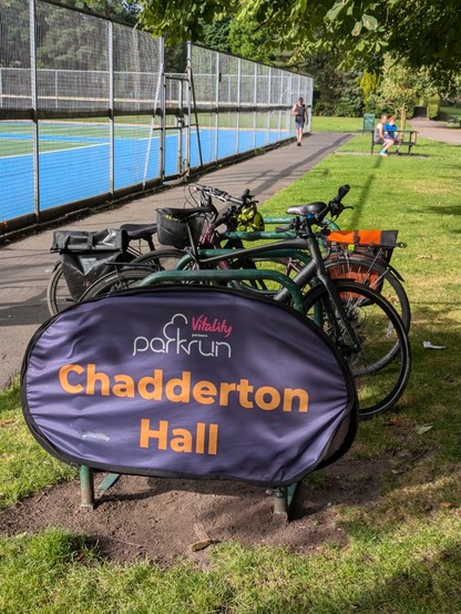 The oval Chadderton Hall parkrun sign in orange text on a purple background, slotted over a bike rack rung. There are bikes on the rungs behind. In the background grass spreads out to the right, with a couple of people sat at the picnic table in the distance. A path to the left separates the grass from the fence enclosing a tennis court with a blue and red surface. Mature horse chestnut trees provide a little shade from the morning sun.