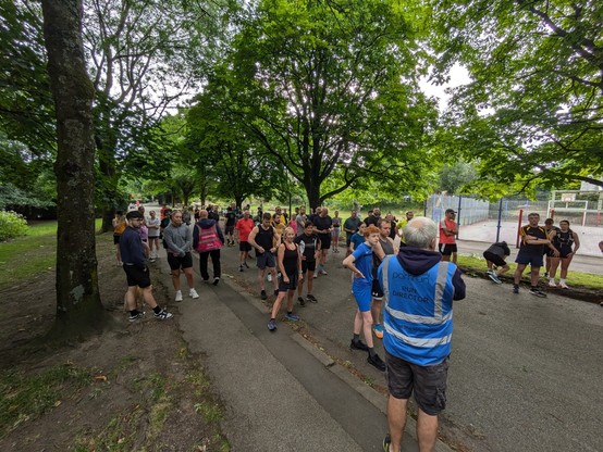 Runners assembled on a wide tarmac path listening to the pre-run speech from the run director who is wearing a blue high visibility tabard. Trees line the sides of the path.