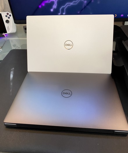 Two Dell laptops, one silver and one white, are placed on a desk. A gaming console controller is visible in the background.