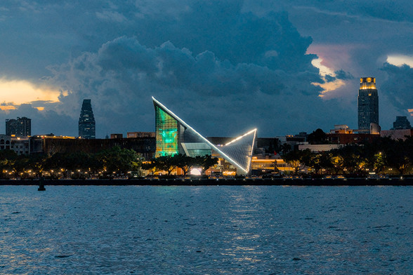 A waterfront cityscape at dusk featuring a modern angular building illuminated with lights and tall buildings in the background under a cloudy sky.