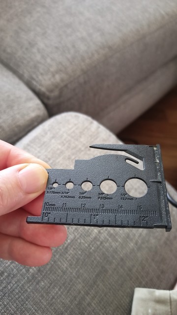 My hand, holding a credit card sized bit of shaped plastic with different sizes holes and markings showing the hole sizes and a ruler at the bottom in mm and inches. At the top there is a hook shaped cut out.