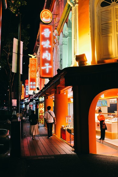 Outside a Chinese pork jerky store with orange pilors, walls and neon signs.  A salesgirl in orange and black uniform too.