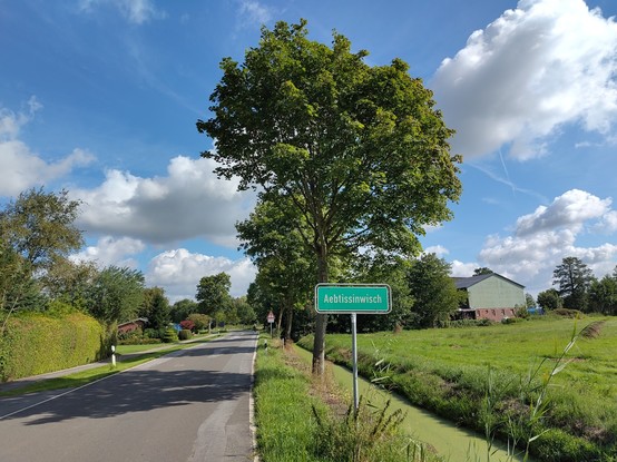 Blick entlang einer Landstraße. Ein Ortsschild mit dem Namen 