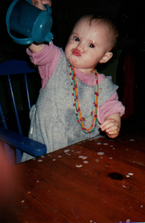 A young, petulant infant seated in a blue high chair wearing a blue & pink outfit and a beaded necklace upturning a sippy cup full of milk on her head and on a wooden table. She has a stern & pouty expression upon her face.