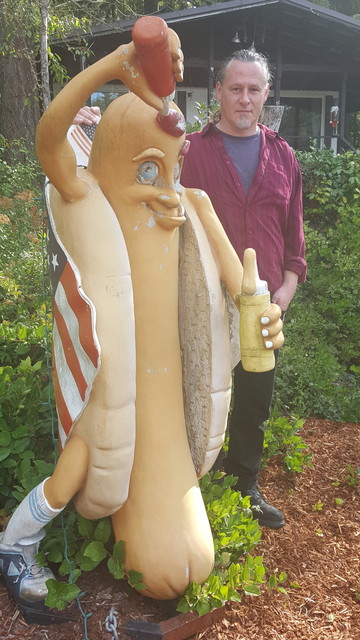 A picture a a man with a red button down shirt & a blue t-shirt standing next to a life-sized statue of a hot dog in a bun draped in an American flag pouring a bottle of ketchup over his head.