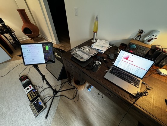 View of a tastefully appointed basement office where we see a desk with a laptop and various other music related to Raitis, and then on the floor a series of guitar pedals, an iPad floating on a microphone stand and a Bogner guitar cabinet tilted up towards the player