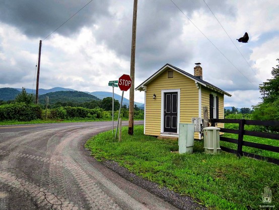 Small tidy bus stop built like a tiny farmhouse at the intersection of rural roads in the foothills of the blue ridge mountains. A crow flies out of frame. 