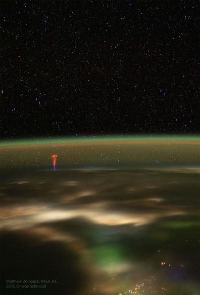 picture of the limb of earth at night with a greenish airglow layer marking earths atmosphere. In the center, the gigantic jet  - a collumn of light, starting in a deep blue color and transitioning to a deep red color rises up from the surface. it branches out towards the top.
City lights from Indonesia are visible in the foreground