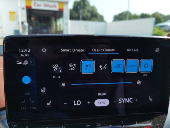 Climate control display of an EV car showing 36C outside temperature and the a/c on max. In the distance outside part of a gas station is visible.