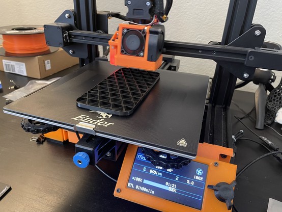 Black and orange Ender 3D printer, half way through printing a black mock smartphone