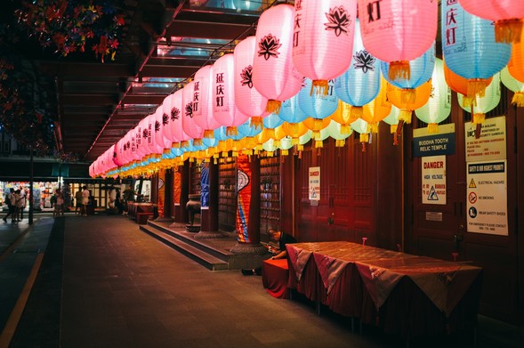 Rows of colorful lanterns decorating the front of a Chinese temple.  There was a man seated checking his phone.