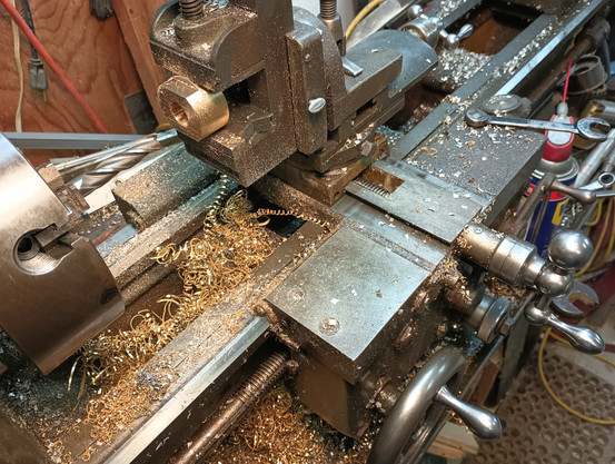 Color photo of a round brass object clamped in a vice that is mounted to the carriage of a metal cutting lathe. There's several handles on the right for controlling how the object is cut. A round drill like tool is mounted in the chuck. For people who have done machine work, this is a mill vise setup on a lathe. Lots of curly twisted gold colored brass turnings that were cut from the object are in the pan area of the lathe.  