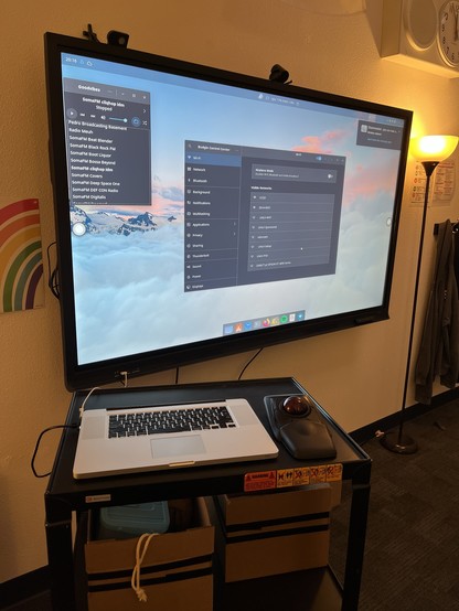 A large display screen showing a desktop interface with various applications and network settings. Below it, a headless MacBook and a mouse are placed on a black table, alongside several storage boxes. The setting appears to be a tech workspace or classroom.