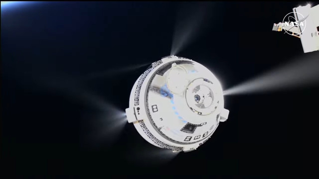 Screenshot of Starliner moving way from the ISS, small thrusters firing