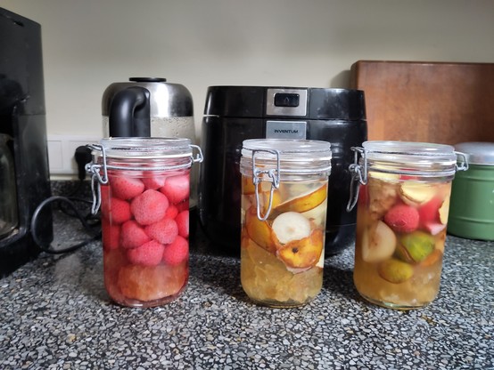 Three mason jars on a counter top. From left to right: filled with strawberries, filled with pears and filled with mixed fruit. The bottom fifth of all jars is filled with rock sugar. In the background, an electric kettle, a rice cooker, a wooden chopping board and an ancient tea kitty.