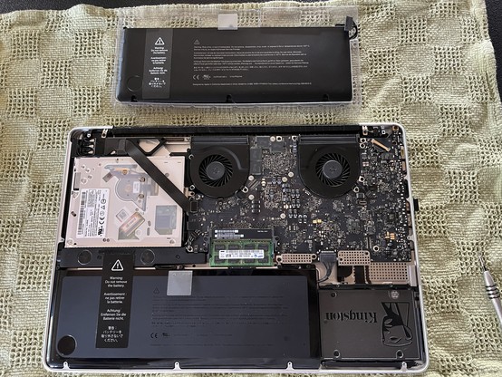 An opened laptop with its components exposed. The new battery placed on top of the laptop. Inside, there are two cooling fans, a logic board, the old battery and a memory module visible. A screwdriver is positioned nearby.