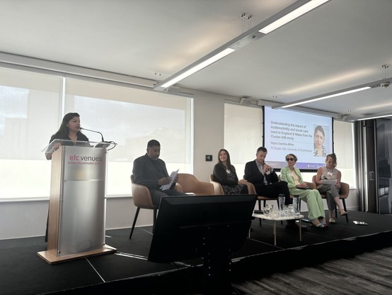 A speaker - Hajira - stands at a podium addressing an audience during a panel discussion. Four panelists are seated, engaging in a discussion about the impact of multimorbidity and social care needs in England and Wales. A presentation is visible in the background.