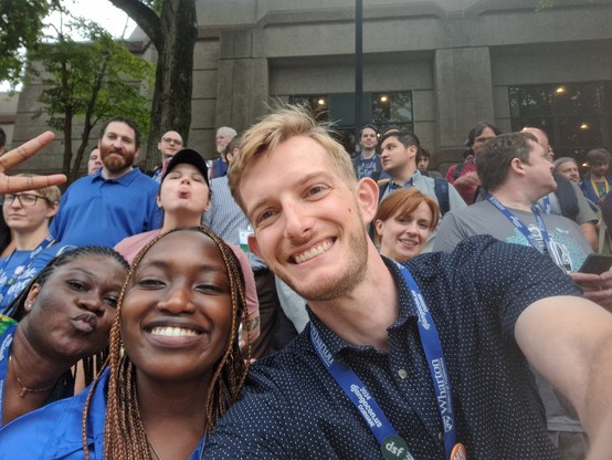 A group of several people. In the forefront there's a selfie of three people. Behind them are several people smiling or looking off in the distance. There's one person comically sticking out their tongue. 