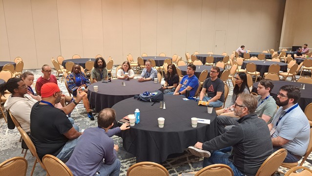 A group of people sitting around two circular tables. The roo. Is large with several tables in the background. Everyone is attentively listening to a single person speaking. 