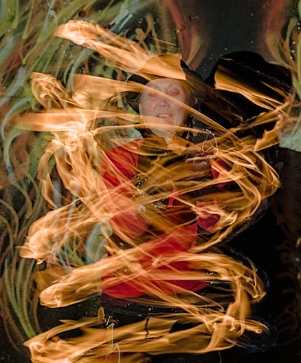 A color portrait photo of a woman with a black derby style hat, red top, and black pants shown slightly below the waist. In the background there is a wall painted with stylized flames in yellow, orange, and white. The woman is spinning poi, little flaming balls on the end of short chains. The long exposure show interlacing flames encircling her body in a corkscrew pattern. Her eyes are cast downward.