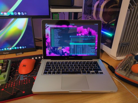 An old MacBook Pro laptop with Linux on it. There are two terminals open on screen, one building a rust program, and the other one showing htop with all cores close to 100% usage.

In the background part of my desk is visible.