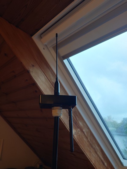 A box with an antenna sticking out of the top vertically upwards, and downwards another to the side, the whole lot on the end of a rod, with a velux window in the background. 