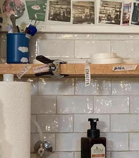 Photo of a sink area with white tile, polaroid photos, tape rolls, paper towels and other work supplies.  Reusable hand-written tape labels indicate coffee regions 