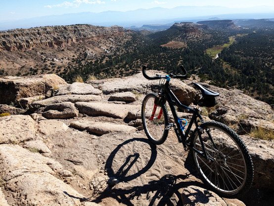 A mountain bike on a rocky mesa edge, looking out into the canyons and finger mesas nearby. There's a road visible at the bottom of the nearest canyon. In the distance, the mountains and far mesas are barely visible in thick haze.