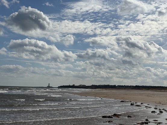 A sandy beach under white and blue skies, with an obscured lighthouse in the far distance. 