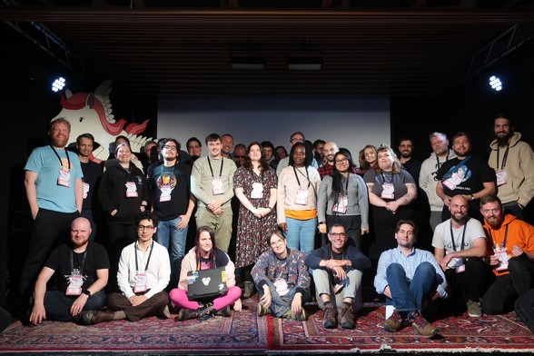 A group photo of conference participants standing on stage in front of the projection screen. A large white and pink pony is visible in the background just behind the participants.