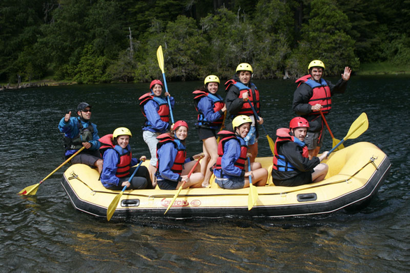 A rafting boat with 9 people. 5 people seated and 4 standing. All of them waving to the camera.