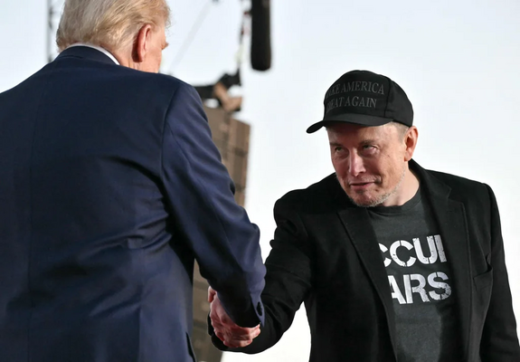 The image shows Musk sharing hands with Trump at a campaign rally. Trump is wearing a dark suit and Musk in a black cap and shirt with text reading 