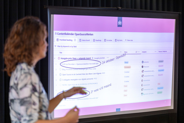 Photograph of a presentation at FOSS4G-BE+NL. On the left there is a female presenter, on the right is the presentation screen showing one of the slides.