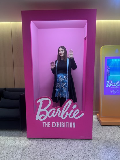 A photo of me, a white woman with long brown hair in a black top, light blue skirt, and black and light blue coat, pretending to be trapped in a life-size Barbie box