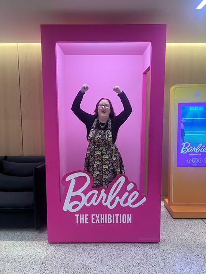 A photo of Clare, a white woman with red hair in a black top and colourful dungarees, pretending to be trapped in a life-size Barbie box