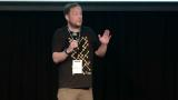 A still of David standing on stage in Puppet t shirt with microphone giving his talk