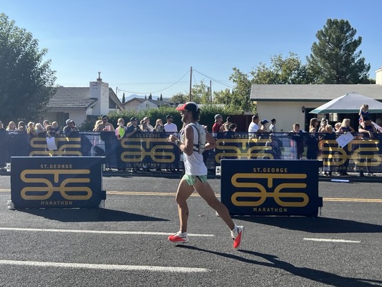 Photo of my in stride running toward the finish line at the St. George Marathon. A crowd of spectators behind a fence on the side of the street is visible in the photo.