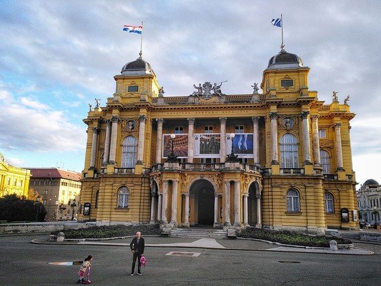 The wind picking up the flags, the light is just right, as a little super-girl strolls past the puddle with her grandpa, creating a theatrical scene just outside the Croatian National Theatre.