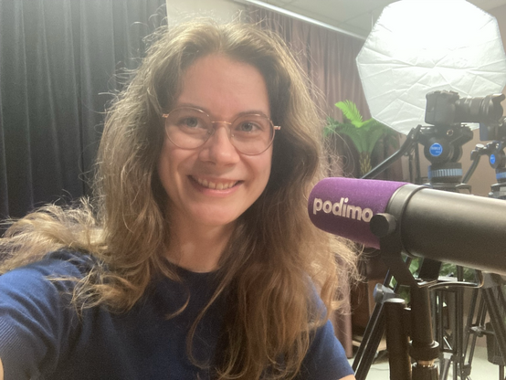 A photo of Judith van Stegeren in a podcasting studio. There are cameras and studio lights in the background. In front of Judith there’s a microphone with a purple windscreen.