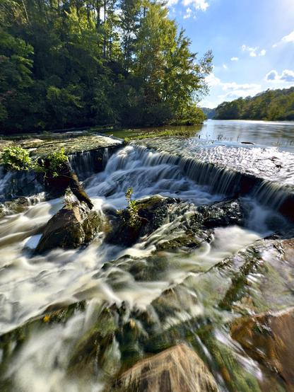 View from below an outflow over a small dam, looking up a winding lake through rolling hills under a blue sky. Water rushed over tumbled rocks and moss covered logs.