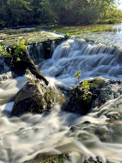 Outflow over a small dam, looking up a winding lake through rolling hills under a blue sky. Water rushed over tumbled rocks and moss covered logs. The long exposure gives the water a sense of flow and movement.