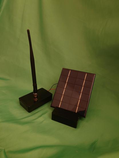 Small black shiny plastic box with large antenna sticking out vertically from the middle of its top. Out of the box run a black and a red cable, into a small shiny solar panel that is angled slightly upwards by being placed in a rubber block with a slot that holds the solar panel at a slant. The background is a green cloth. 