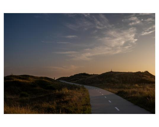 A landscape with a sunset. There’s a cycle path in the landscape, and a cyclist can be seen at the back of the landscape